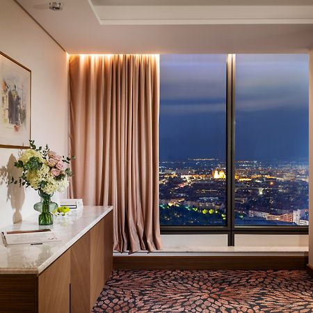 Grand Hotel Millennium Sofia Bagian luar foto The photo depicts a modern hotel room or a lounge area with a stunning view of a cityscape at night. On the left, there's a wooden console table with a vase of flowers and a bottle of water. A piece of art is framed on the wall above the table. The r