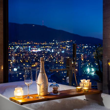 Grand Hotel Millennium Sofia Bagian luar foto The photo features a luxurious bathtub placed by a large window, showcasing a scenic city view at night. There is a wooden tray resting on the edge of the tub, displaying a bottle of champagne, two glass flutes, and several lit candles, creating an a