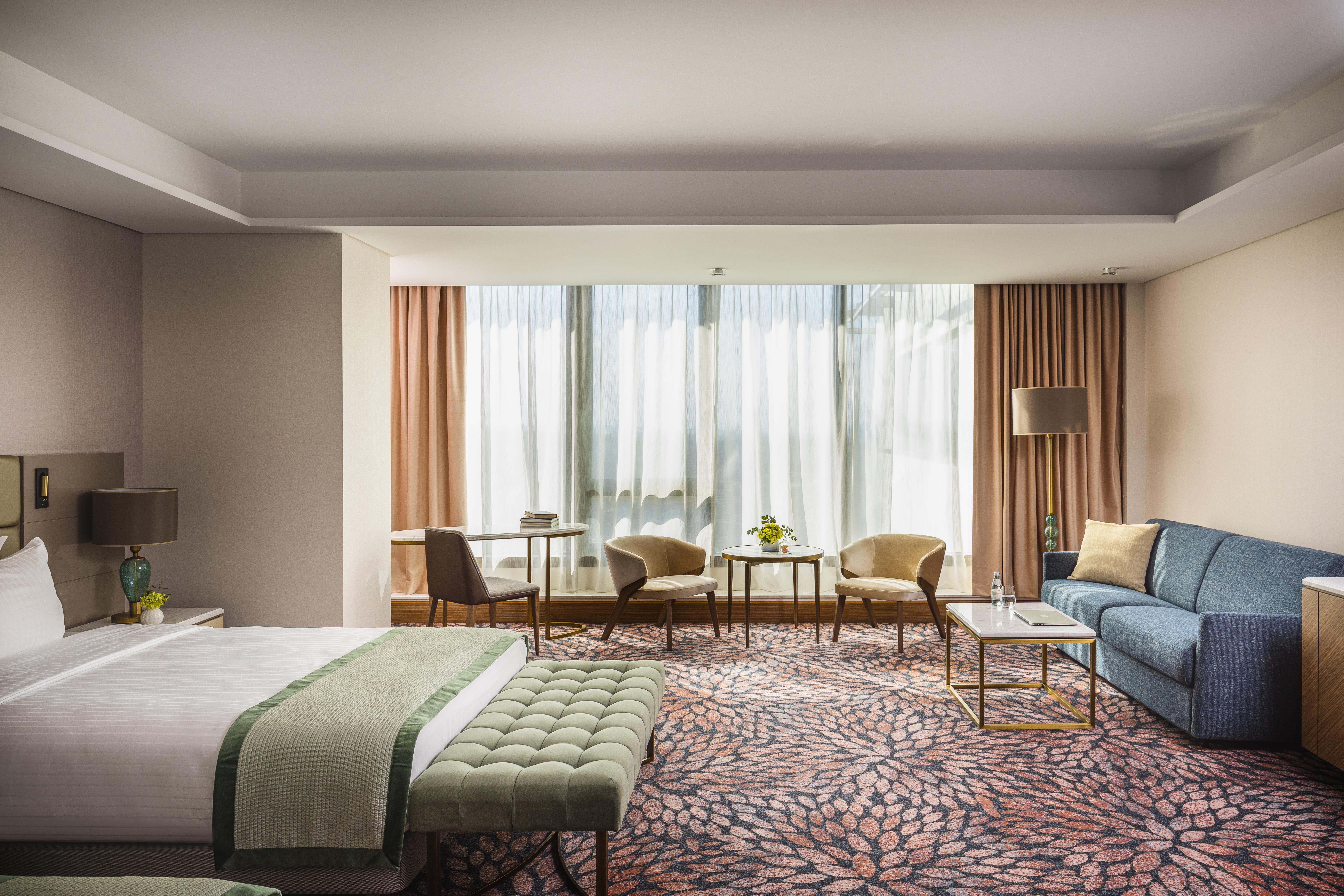 Grand Hotel Millennium Sofia Bagian luar foto The photo shows a modern hotel room featuring a spacious layout. On one side, there is a large bed with a green throw blanket and a tufted bench at its foot. The room is well-lit by large windows dressed with light curtains, which allow natural light