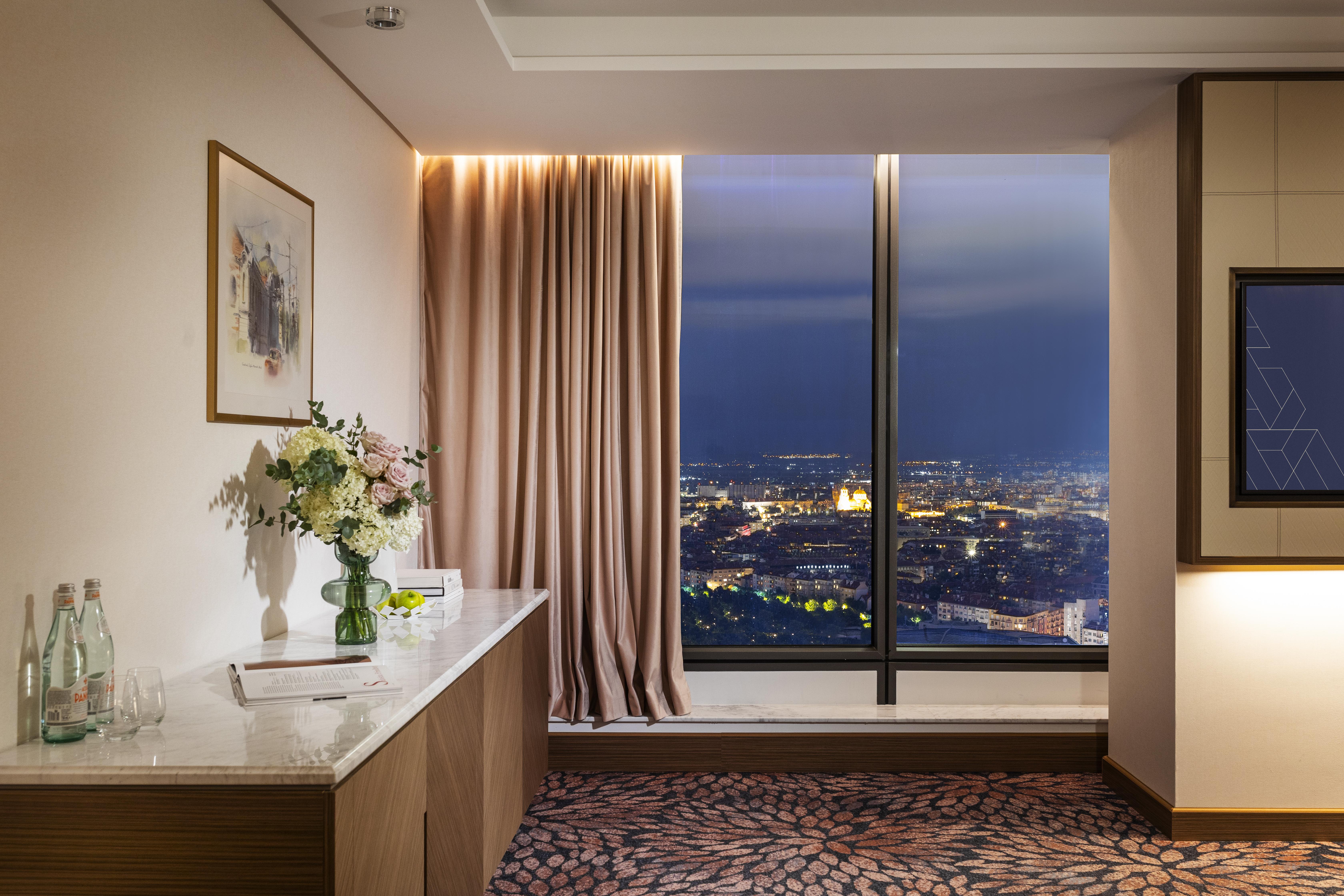 Grand Hotel Millennium Sofia Bagian luar foto The photo depicts a modern hotel room or a lounge area with a stunning view of a cityscape at night. On the left, there's a wooden console table with a vase of flowers and a bottle of water. A piece of art is framed on the wall above the table. The r