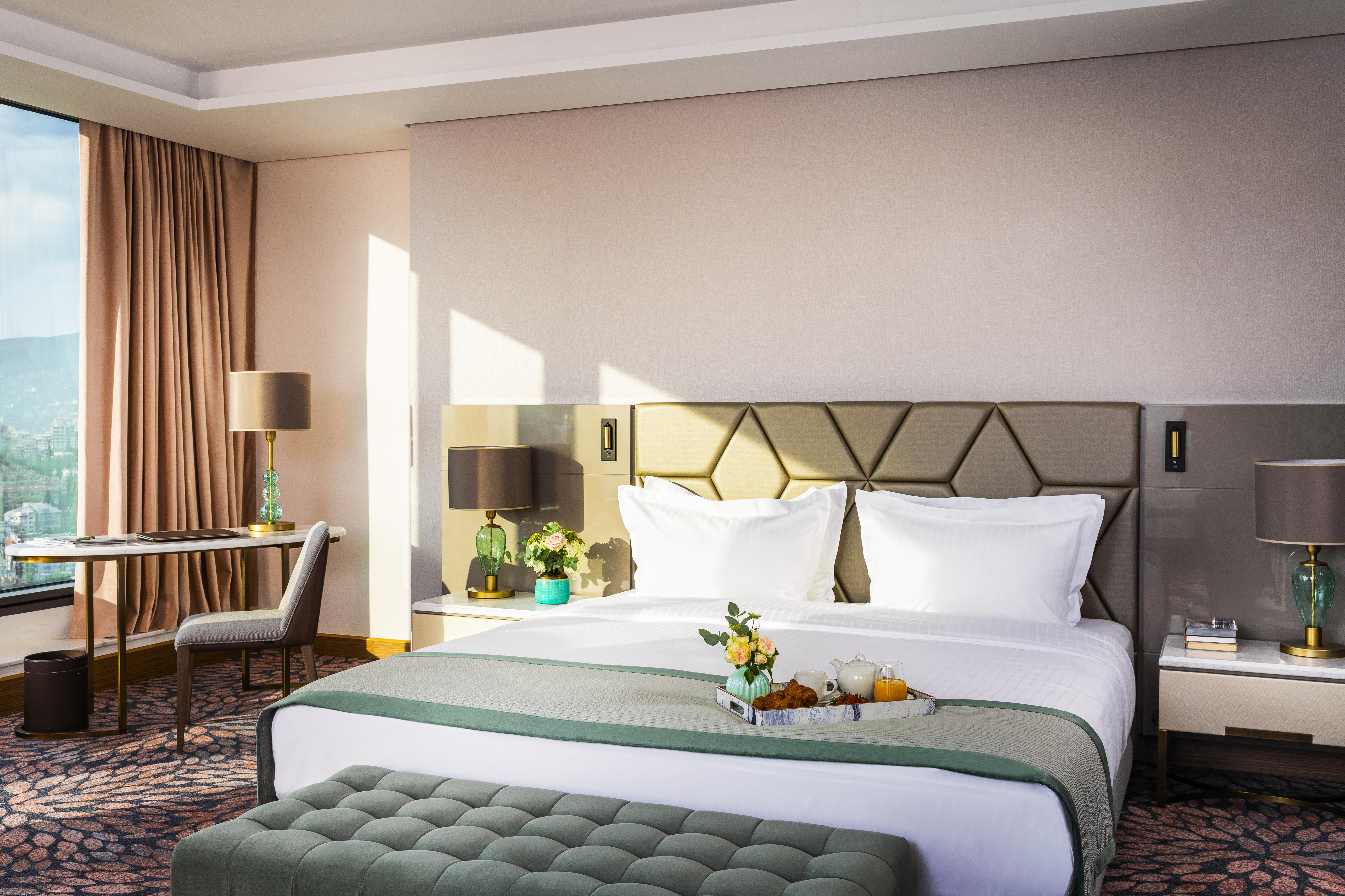 Grand Hotel Millennium Sofia Bagian luar foto The photo depicts a stylish hotel room featuring a large bed with white linens and a green decorative throw. Behind the bed is a geometric-patterned headboard. To the side, there are modern bedside lamps and a small table with a vase of flowers. A se