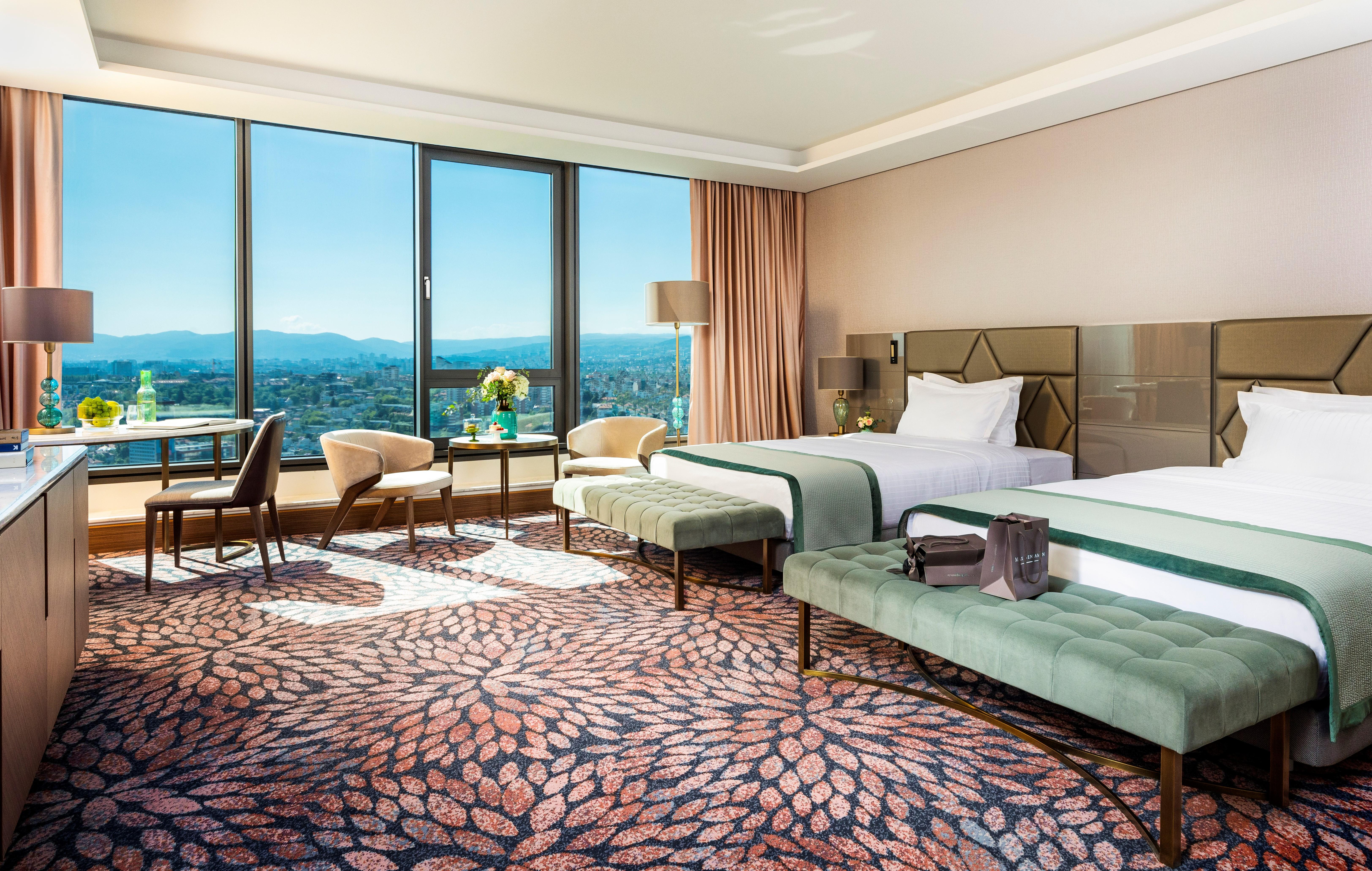 Grand Hotel Millennium Sofia Bagian luar foto The photo depicts a luxurious hotel room featuring two beds with elegant bedding. The room has large windows that offer a panoramic view of the city and mountains in the background. There is a decorative area rug with a floral pattern covering the fl