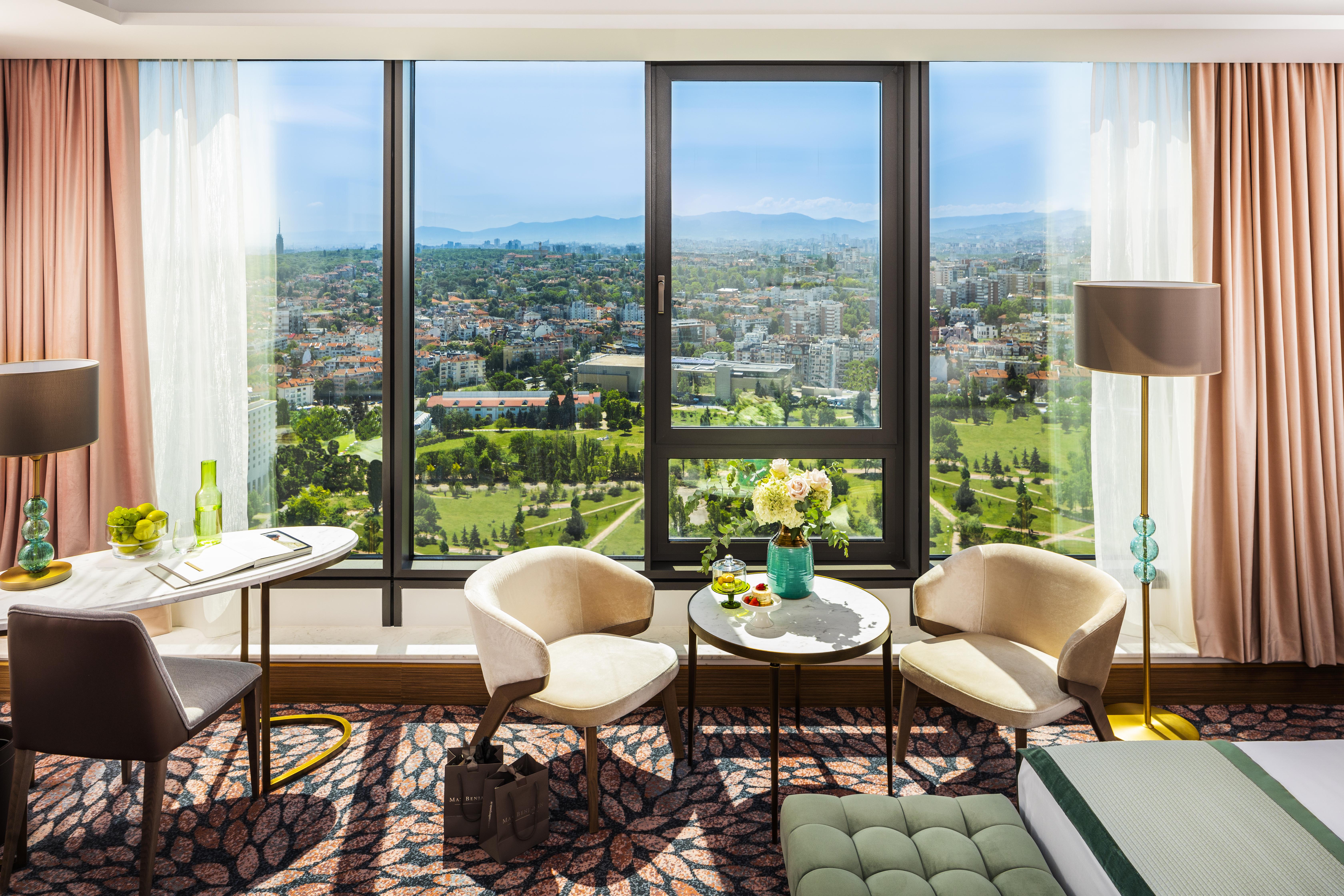 Grand Hotel Millennium Sofia Bagian luar foto The photo shows a stylish hotel room with large windows providing a beautiful view of a cityscape and landscape beyond. There are two modern chairs and a small round table set near the windows, accompanied by a vase of fresh flowers. The room feature