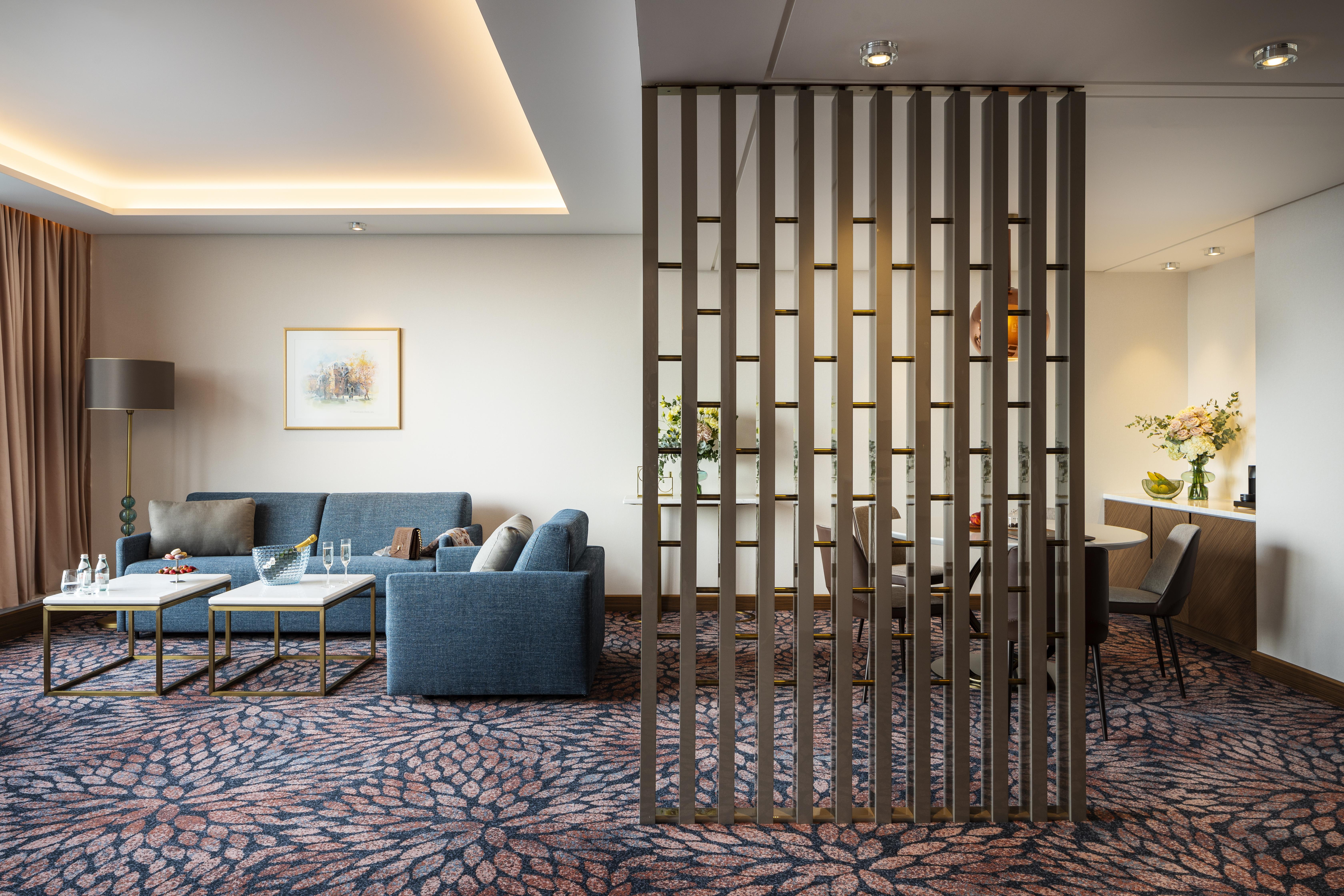 Grand Hotel Millennium Sofia Bagian luar foto The photo shows a modern living space with a comfortable seating area featuring a blue couch and two coffee tables. There is a decorative partition made of vertical wooden slats, which separates this sitting area from a dining space. The dining area 