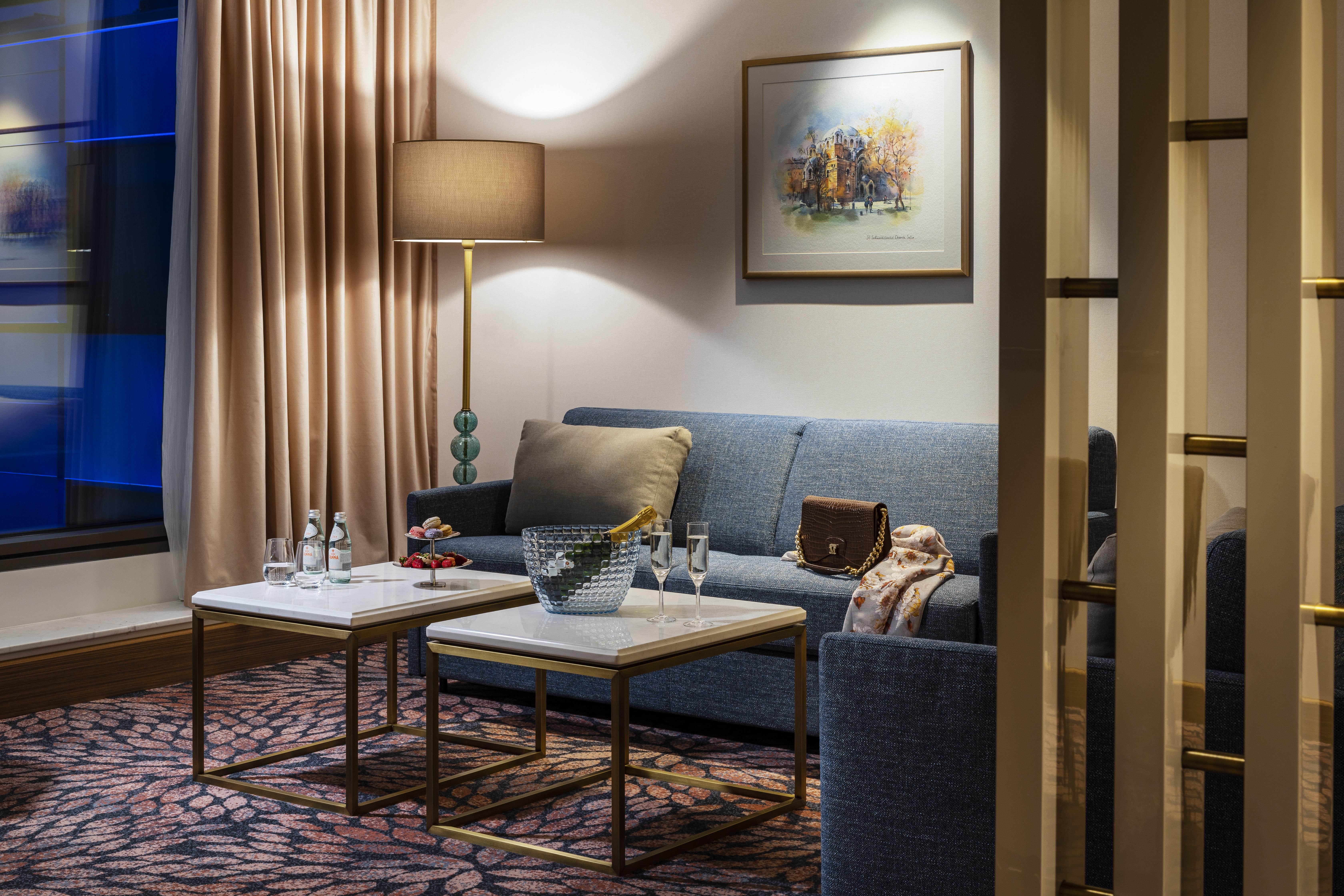 Grand Hotel Millennium Sofia Bagian luar foto The photo shows a cozy and elegantly decorated living space. It features a blue sofa against a wall, complemented by a painting above it. There are two small marble-top coffee tables with gold frames, one of which holds decorative items and glasses. 