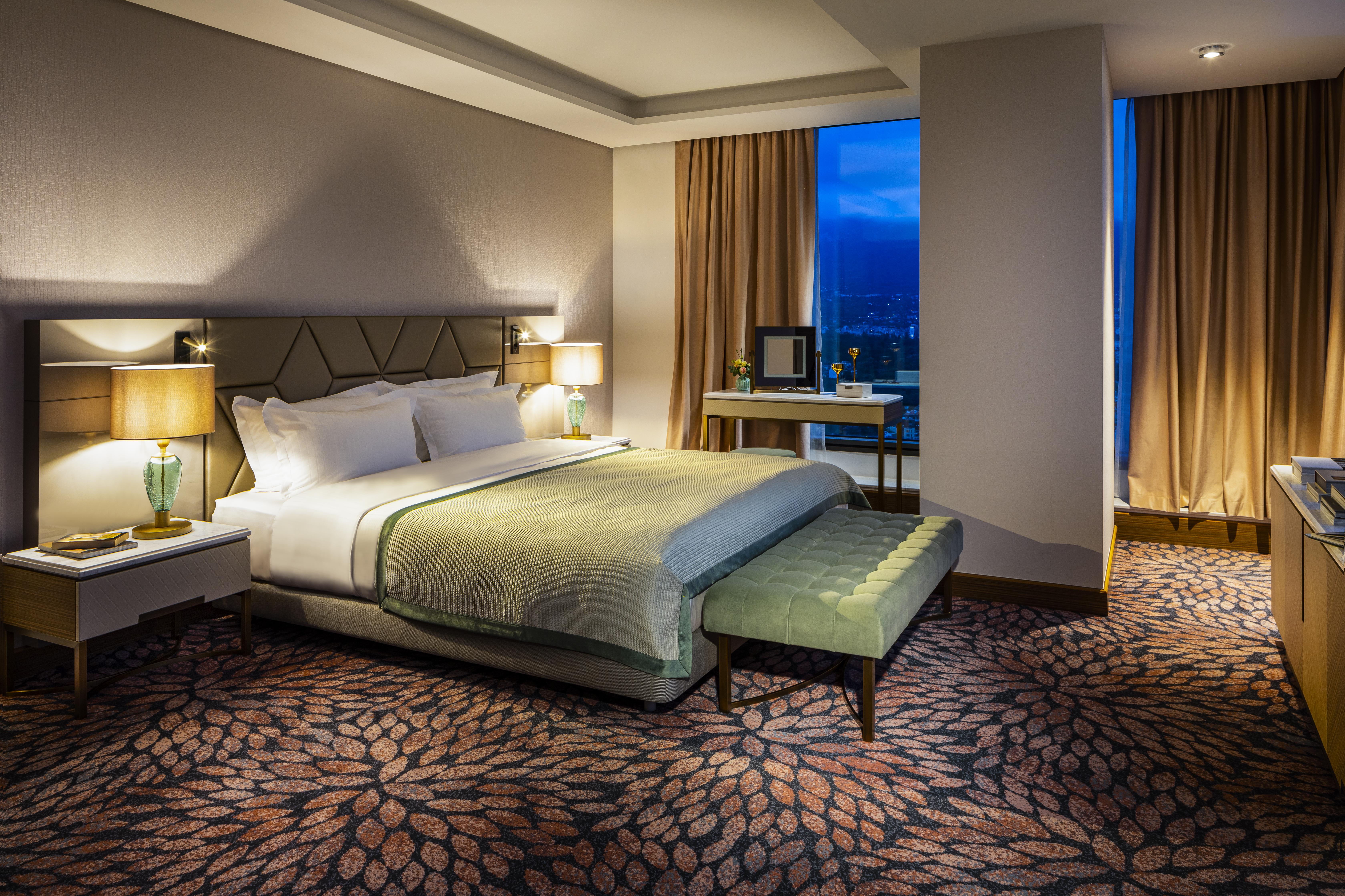 Grand Hotel Millennium Sofia Bagian luar foto The photo shows a modern hotel room featuring a spacious design. It has a large bed with a light green throw blanket positioned in the center. On either side of the bed, there are stylish bedside tables with lamps. 

The floor has a patterned carpet,
