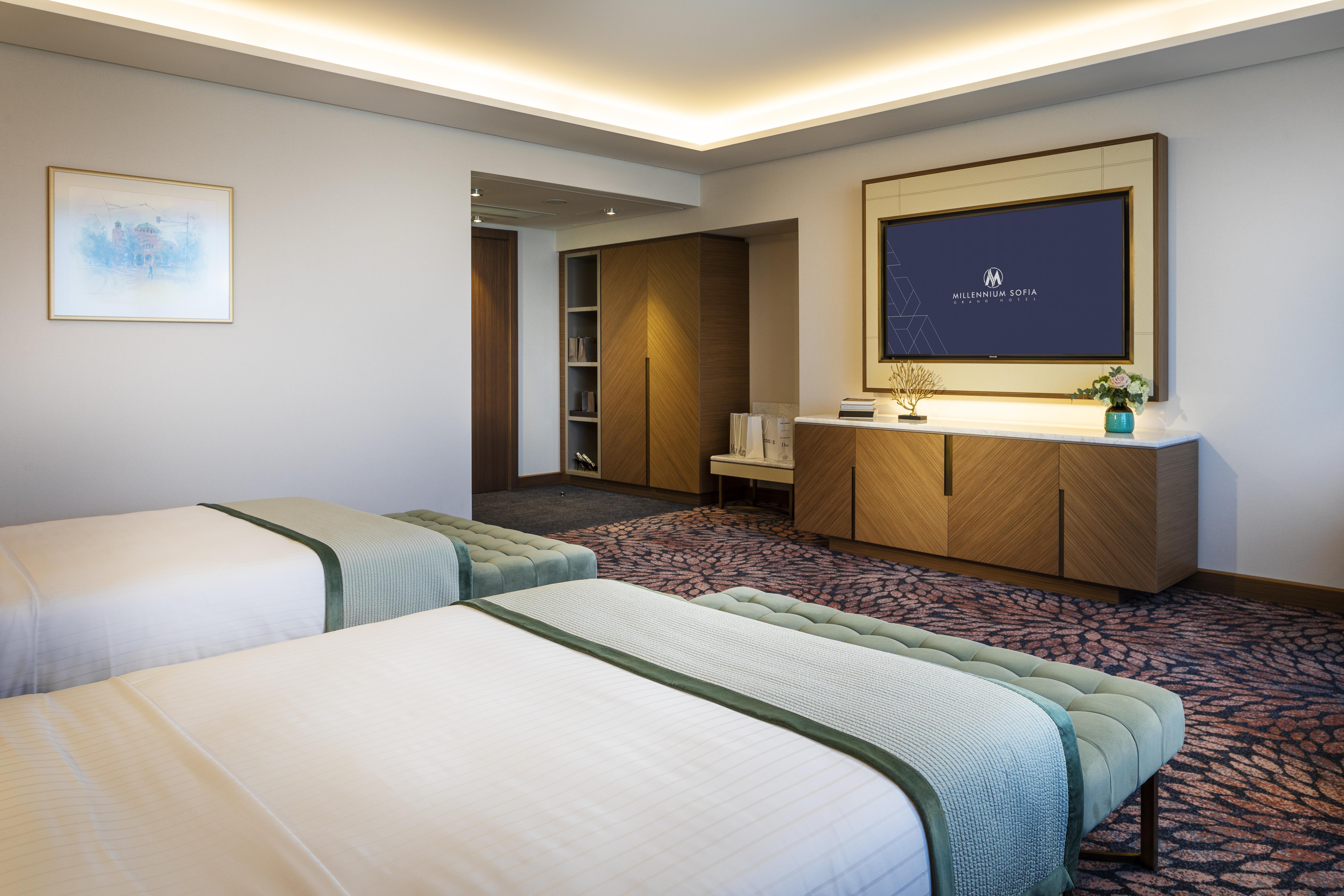 Grand Hotel Millennium Sofia Bagian luar foto The photo shows a modern hotel room featuring two beds with light-colored bedding and decorative throws. The room has a contemporary design, characterized by neutral walls and a plush carpet. A large television is mounted on the wall across from the 