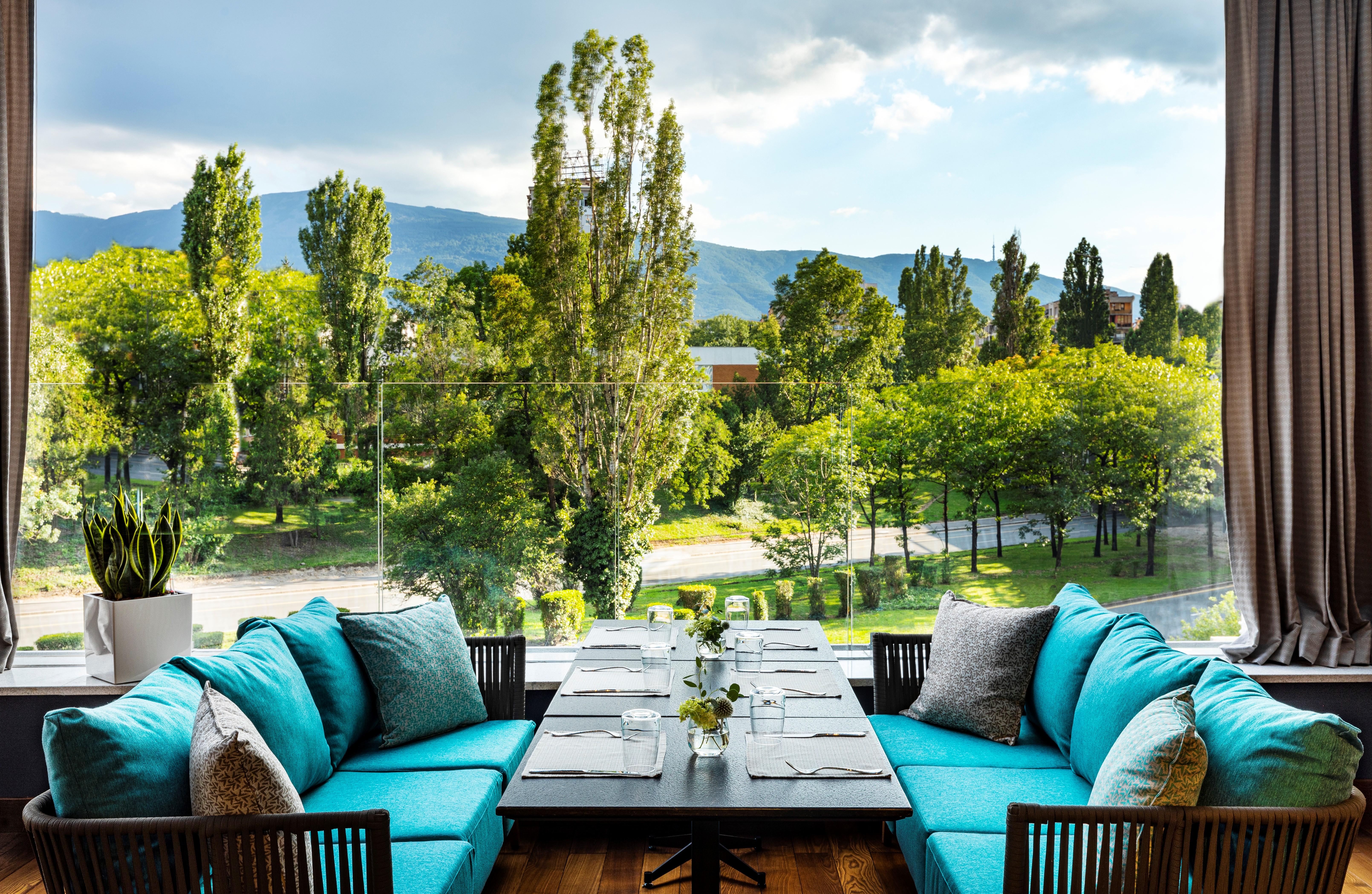 Grand Hotel Millennium Sofia Bagian luar foto The photo shows a bright and inviting dining area with large windows that overlook a lush green landscape. There are several trees in the background, along with a view of distant mountains under a partly cloudy sky. The seating consists of stylish tu