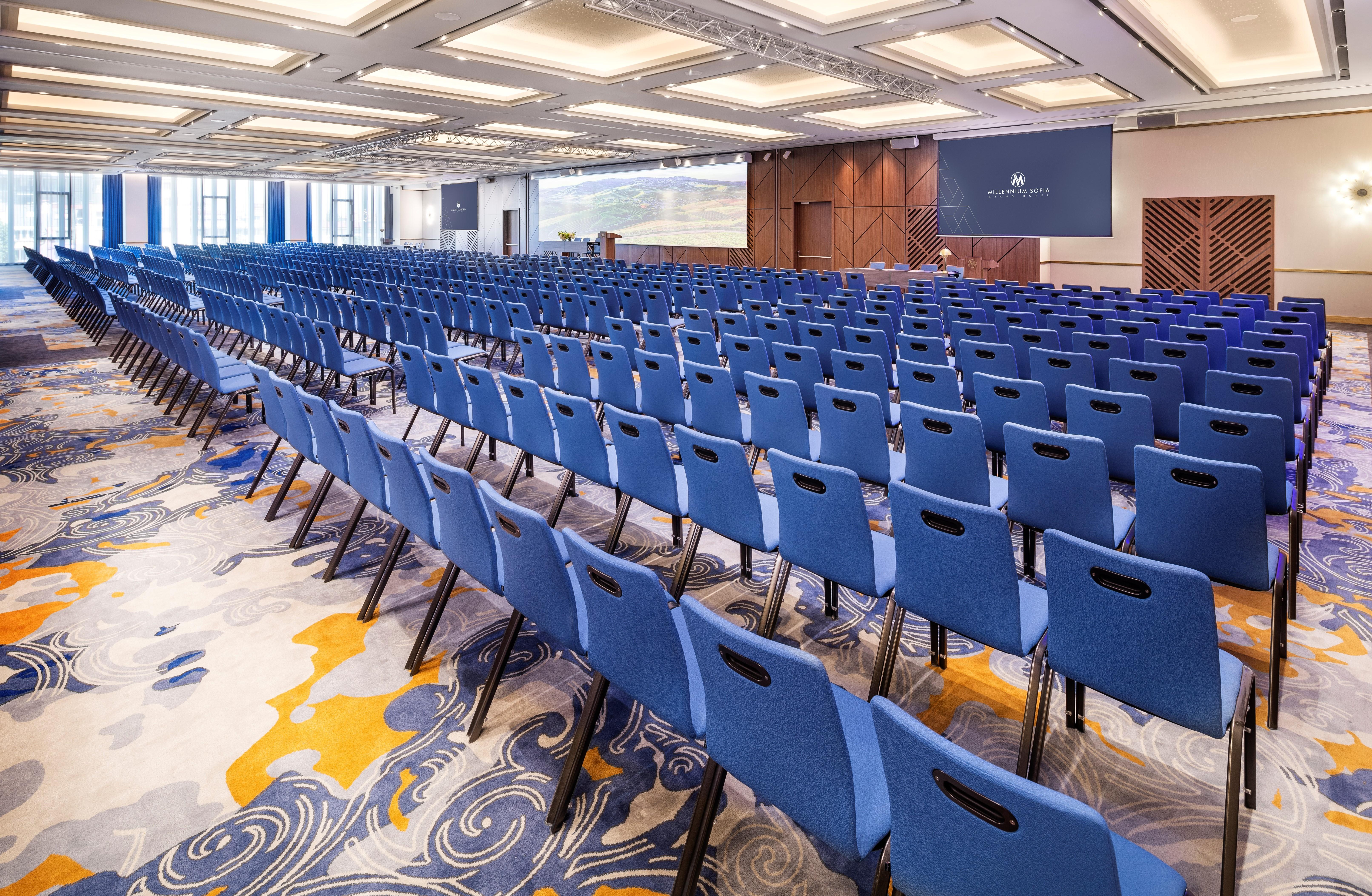 Grand Hotel Millennium Sofia Bagian luar foto The photo shows a large conference or meeting room set up for an event. There are numerous blue chairs arranged in rows, creating a seating area for attendees. The floor features a decorative pattern, and the room has a modern design with ample light