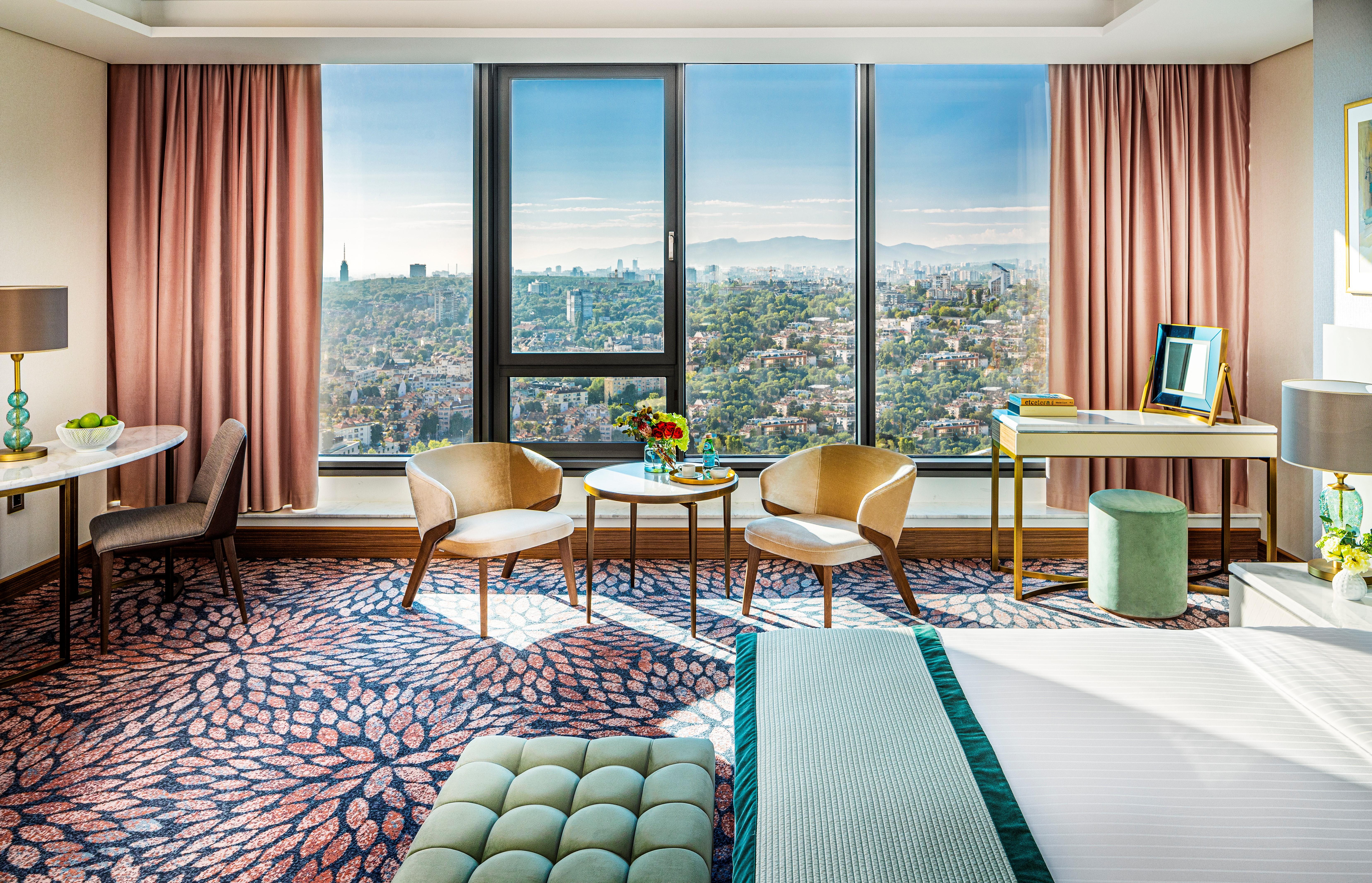 Grand Hotel Millennium Sofia Bagian luar foto The photo shows a stylish hotel room featuring modern decor. There are large windows that offer a panoramic view of the cityscape, allowing natural light to fill the space. The walls are adorned with soft pink curtains. In the room, there are two ele