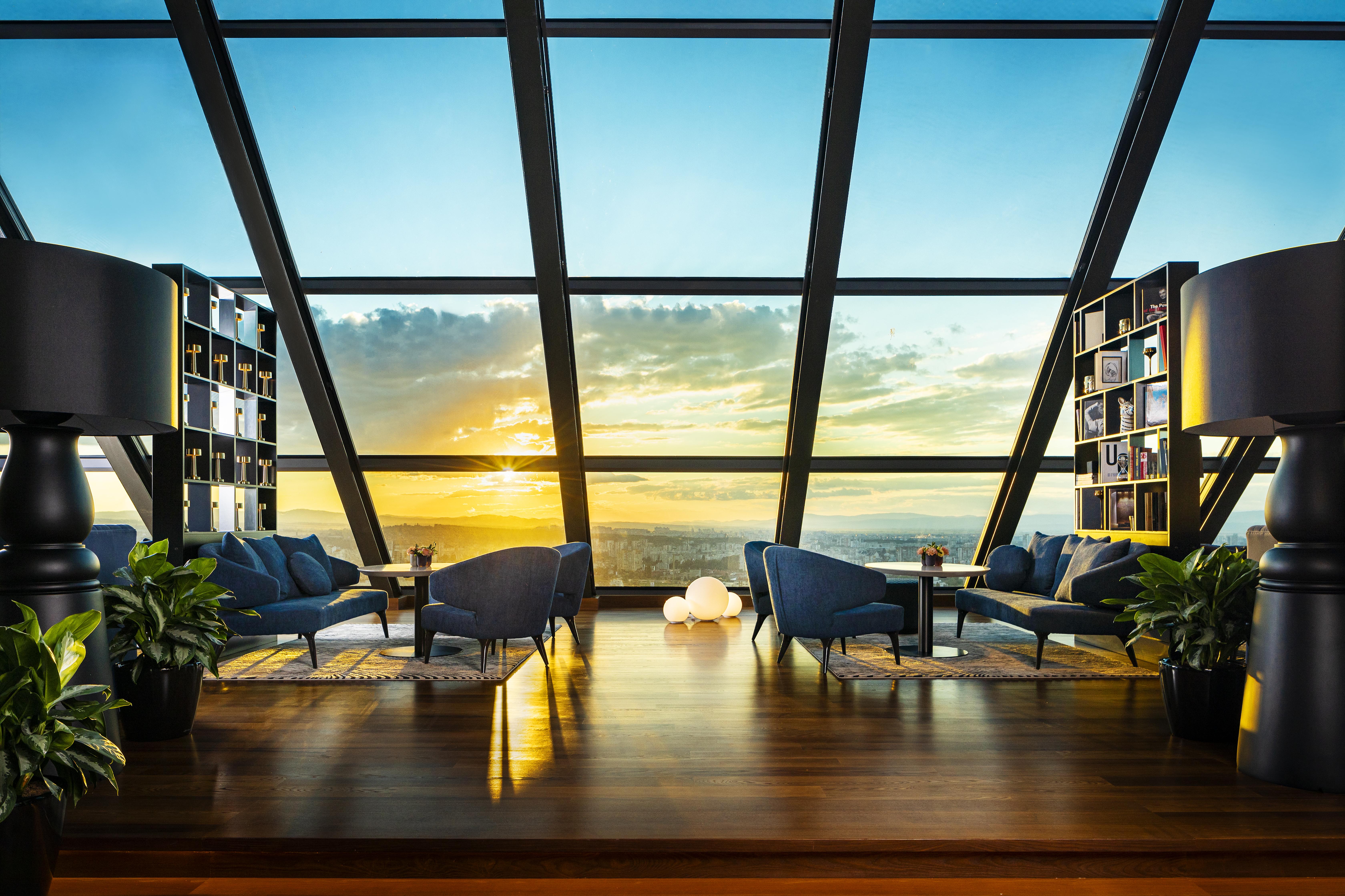 Grand Hotel Millennium Sofia Bagian luar foto The photo depicts a modern, stylish interior of a high-rise space with a panoramic view through large glass windows. The scene features a cozy seating area with several blue armchairs arranged around a low table, creating a comfortable atmosphere. Th