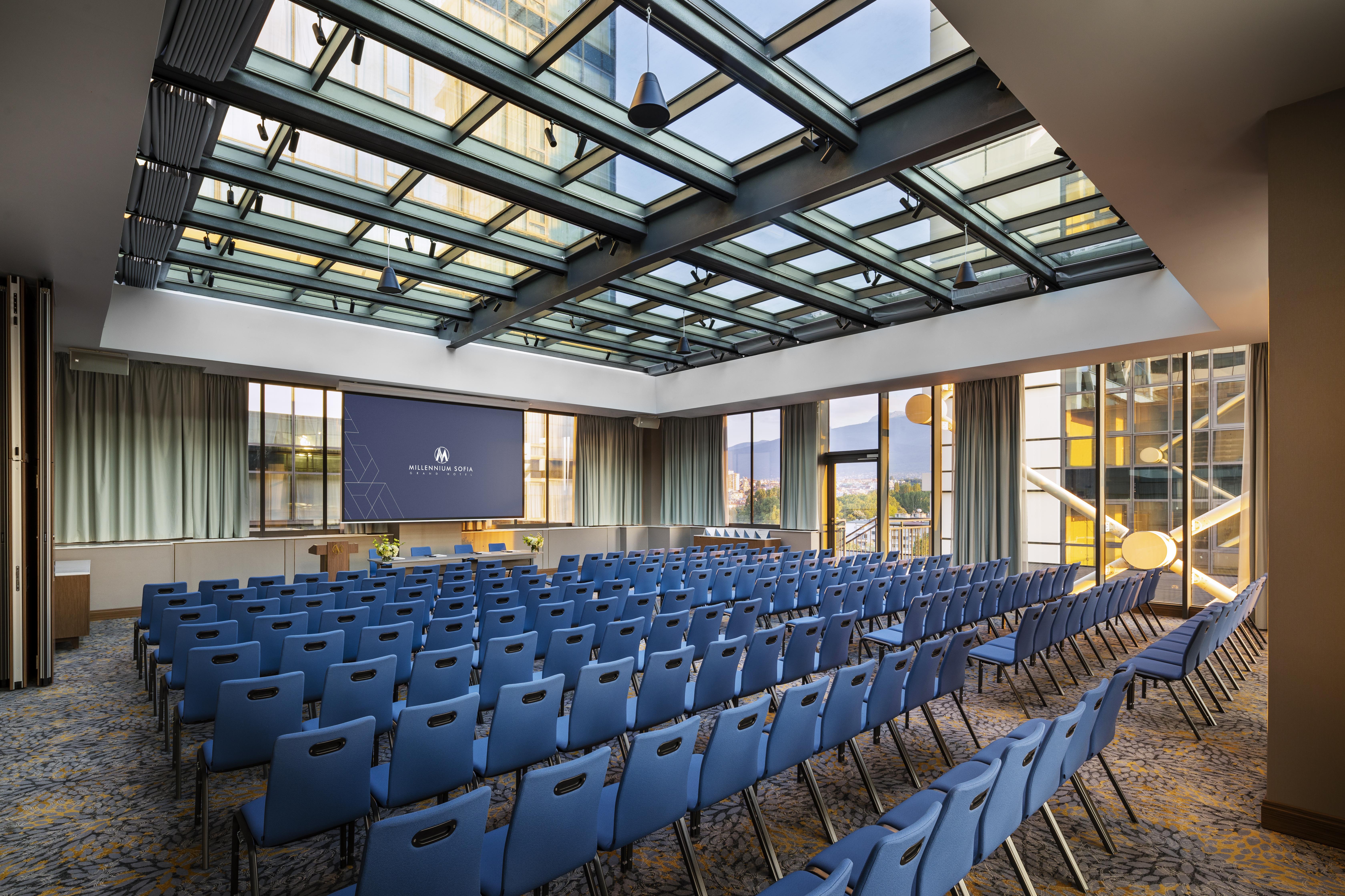 Grand Hotel Millennium Sofia Bagian luar foto The photo shows a large conference room or meeting space. It features rows of blue chairs arranged in a theater style, facing a large screen at the front of the room. The room is well-lit, with a glass ceiling allowing natural light to filter in. The