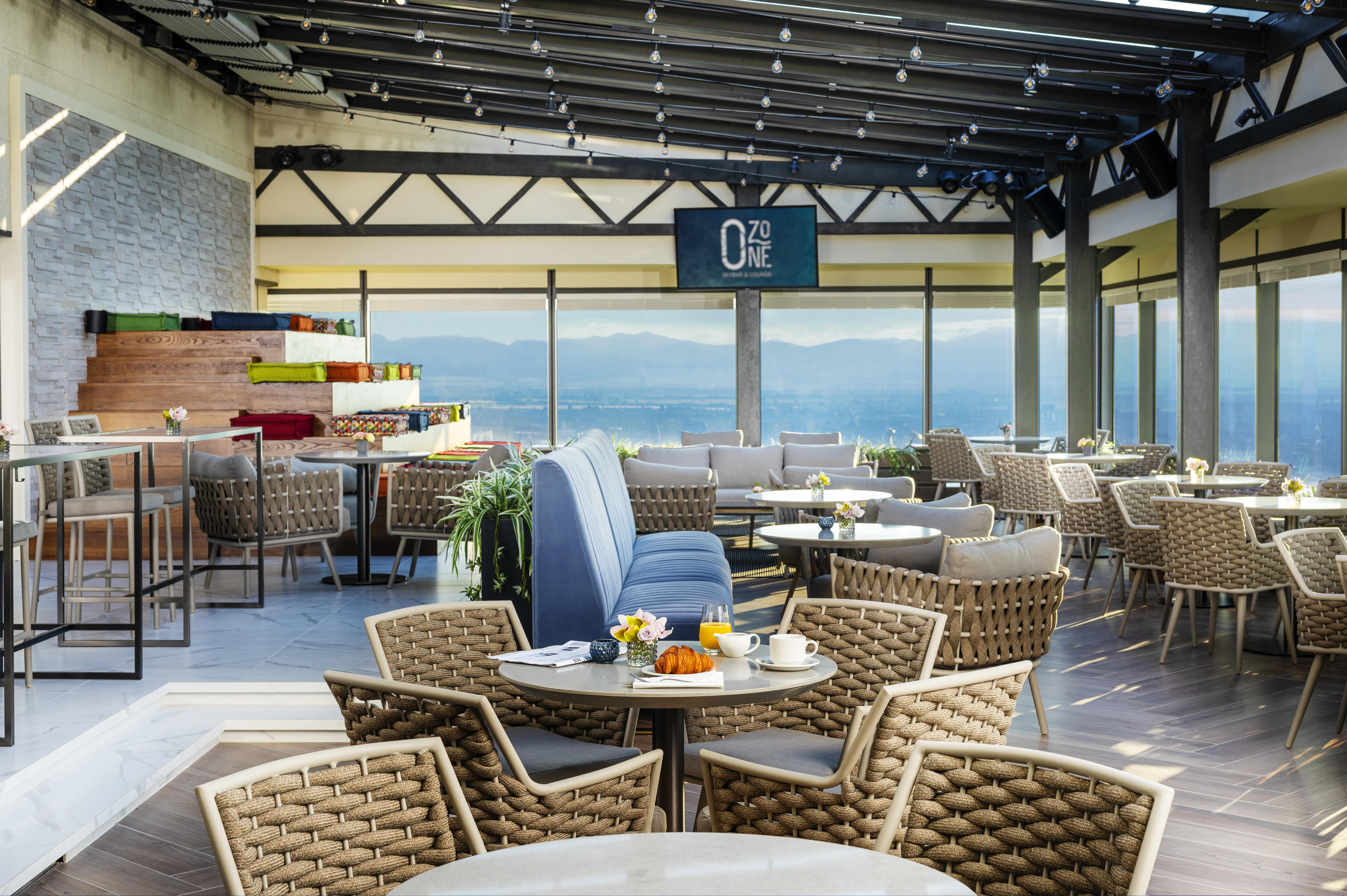 Grand Hotel Millennium Sofia Bagian luar foto The photo shows a modern restaurant or café with a spacious interior. It features a variety of seating arrangements, including round tables surrounded by wicker chairs and lounges. Large windows allow for a panoramic view, which likely includes sceni