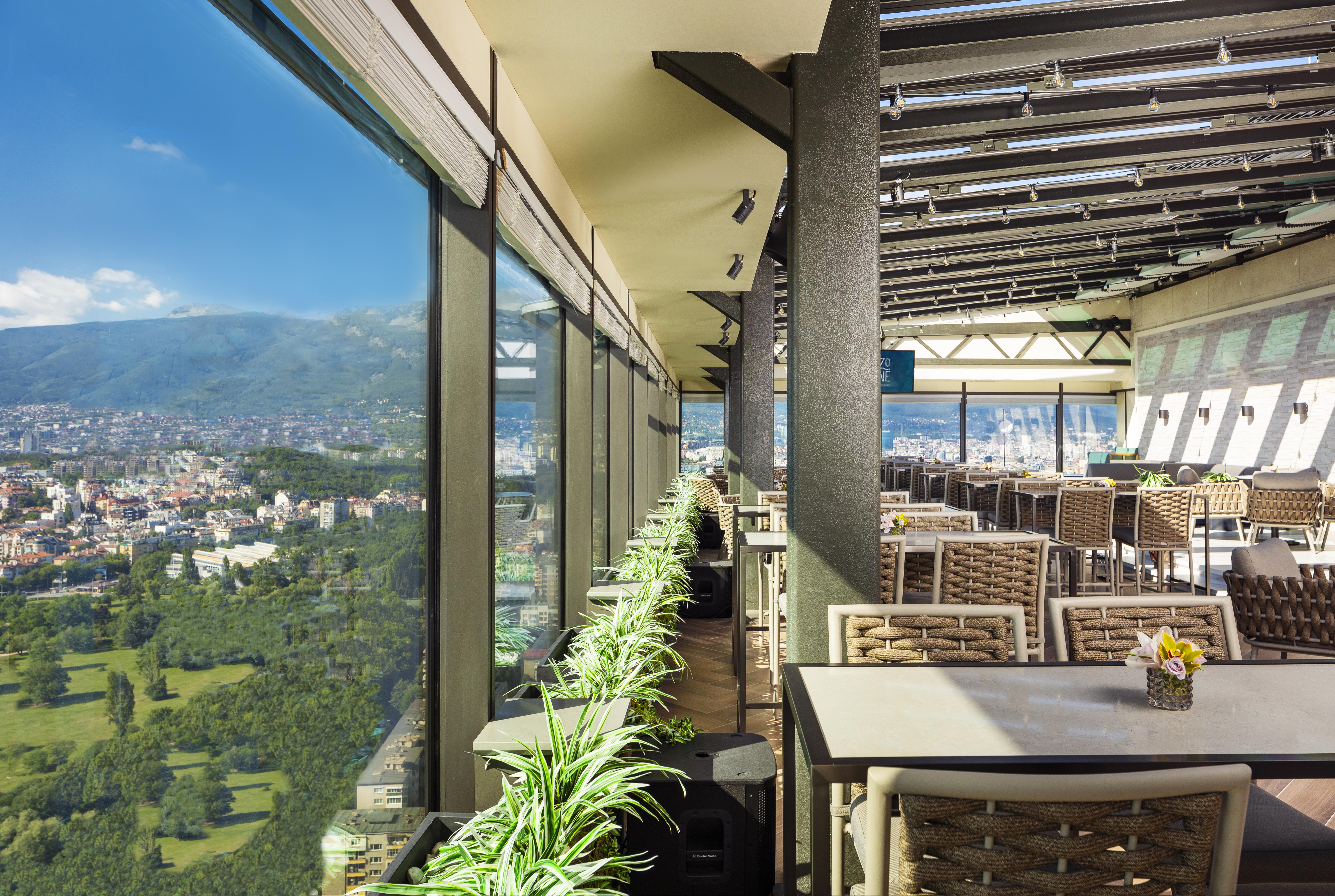 Grand Hotel Millennium Sofia Bagian luar foto The photo shows a modern, spacious restaurant with large glass windows offering a panoramic view of a city and surrounding landscape. The interior features contemporary furniture, including stylish tables and chairs. There are potted plants along the